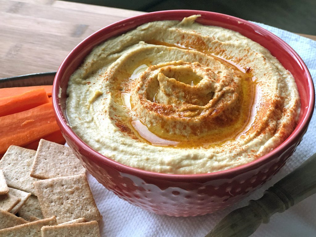 red bowl of garlic hummus with a side of carrot sticks and crackers
