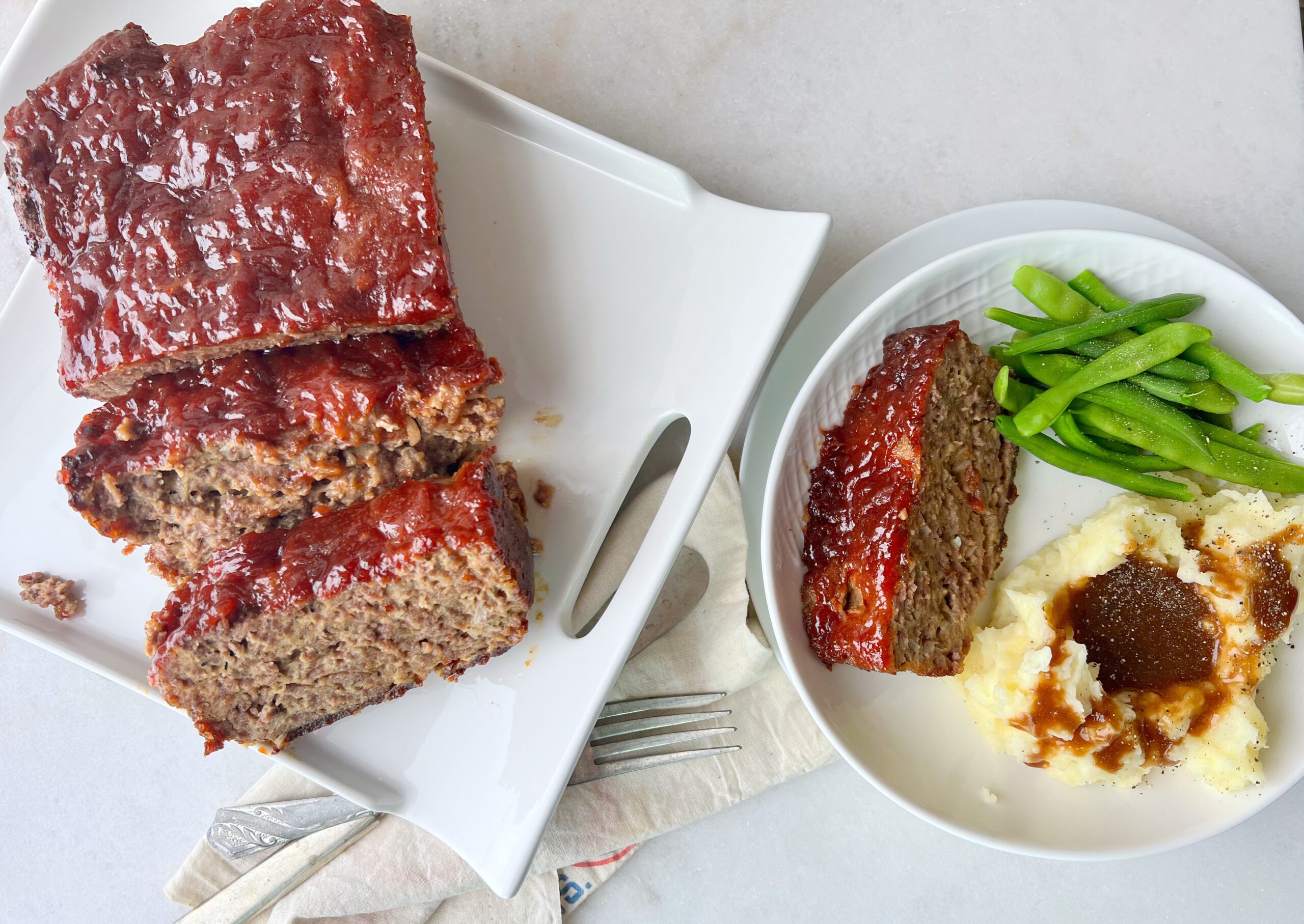 platter with meatloaf and a plate with a slice of meatloaf, mashed potatoes and gravy and green beans