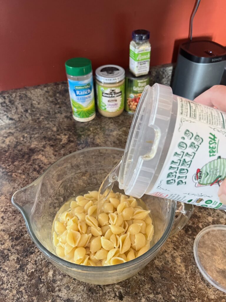 pouring pickle juice into a bowl of cooked pasta shells